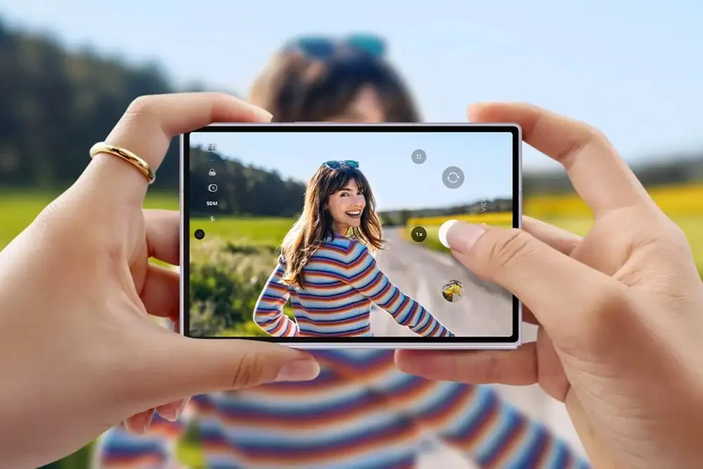 A smartphone camera screen captures a woman in a striped sweater smiling, with on-screen controls including '50M' and zoom options visible.