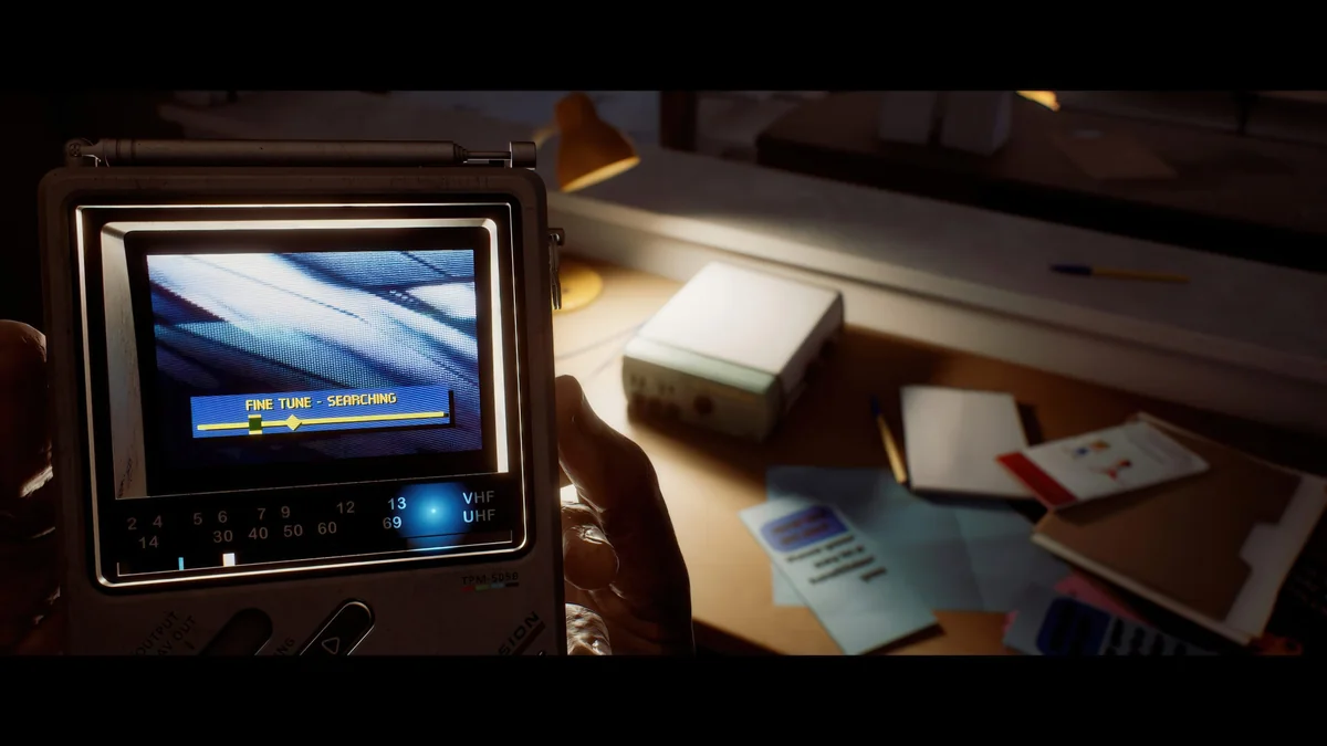 A handheld device labeled TPM-5058 displays 'FINE TUNE - SEARCHING' while a desk in the background contains papers, a pen, and an electronic device.