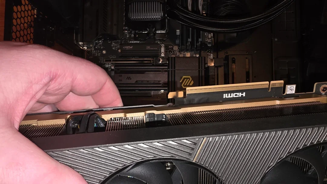A hand is installing a NVIDIA GeForce graphics card into a motherboard inside a computer case, with visible RAM sticks labeled Crucial and a logo on the motherboard.